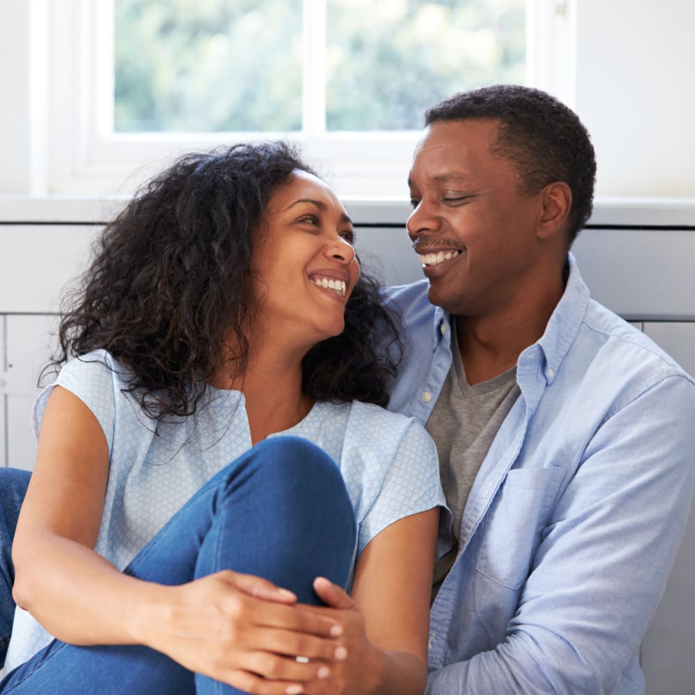 A man and a woman sit next to eachother, looking at eachother and smiling. The moan has her leg up and is holding it with her hands.